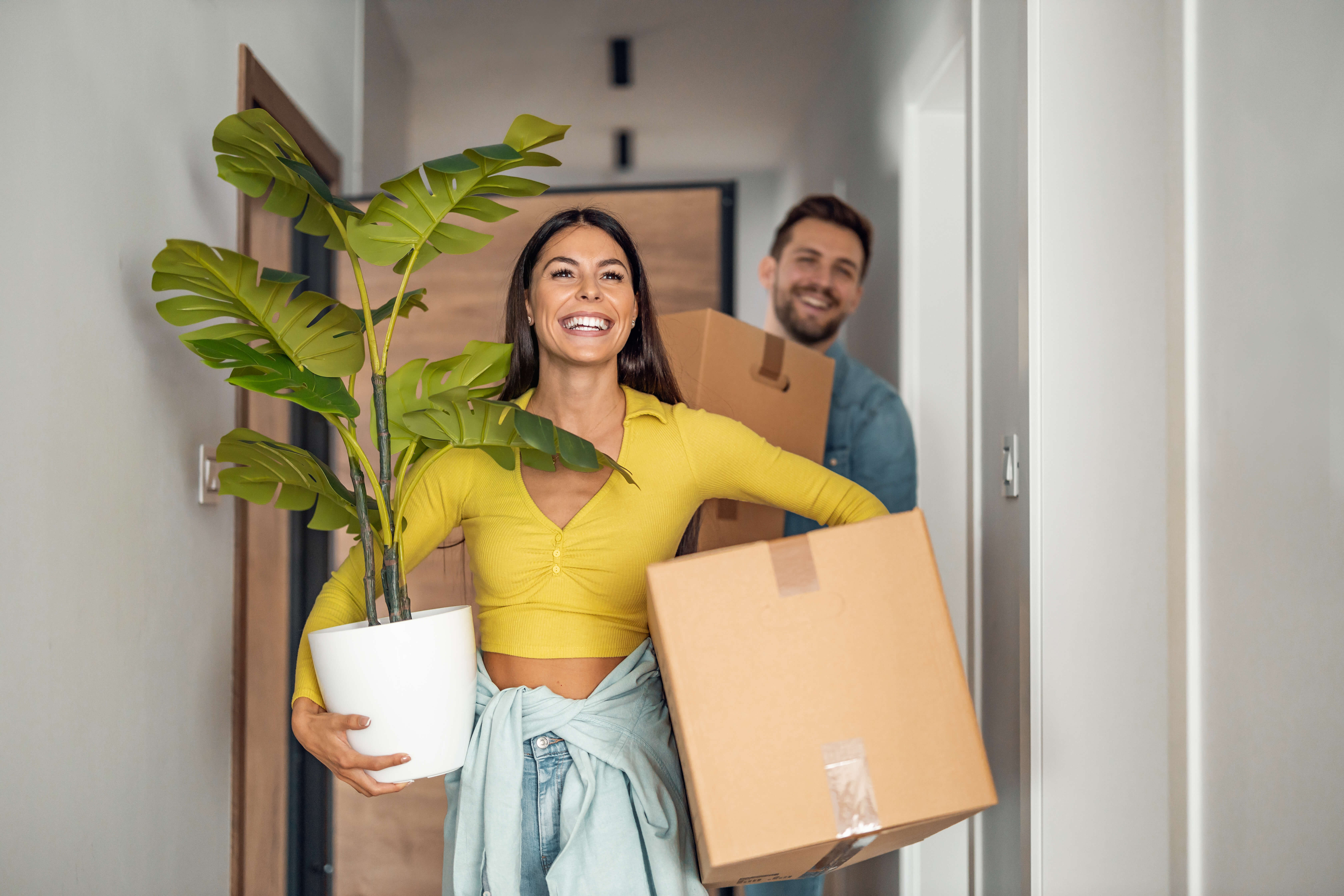 Smiling couple moving into a new home with boxes and plants, representing how finding an agent can help homeowners take advantage of valuable insurance discounts.