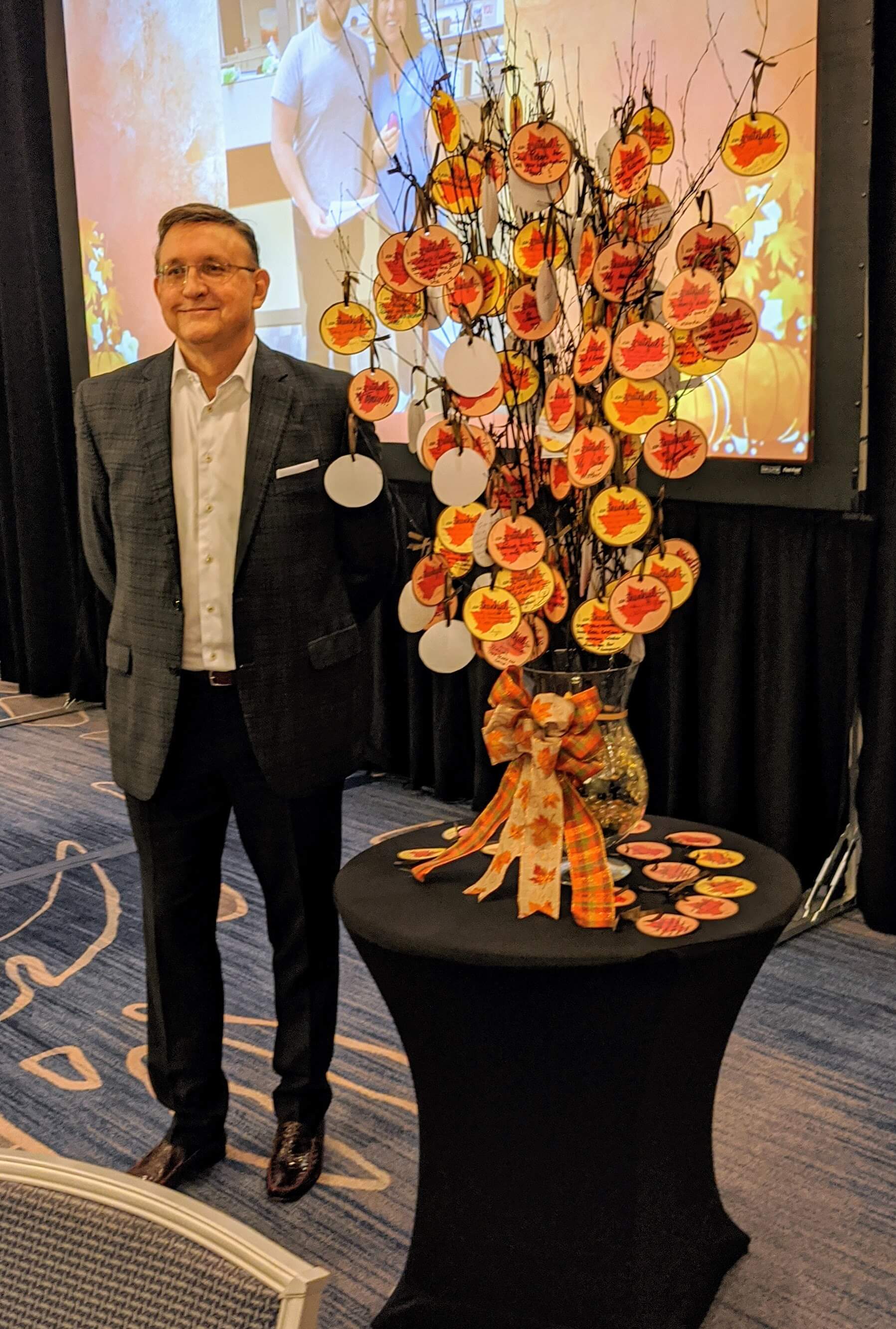 American Integrity CEO Bob Ritchie standing next to a small tree with paper ornaments at the company's annual Gratitude Gathering during Thanksgiving
